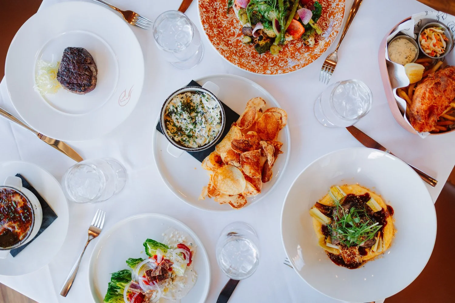 A top-down view of a table with various dishes including a steak, bread with dip, salad, pasta with sauce, and fried chicken with fries, surrounded by glasses of water and cutlery.