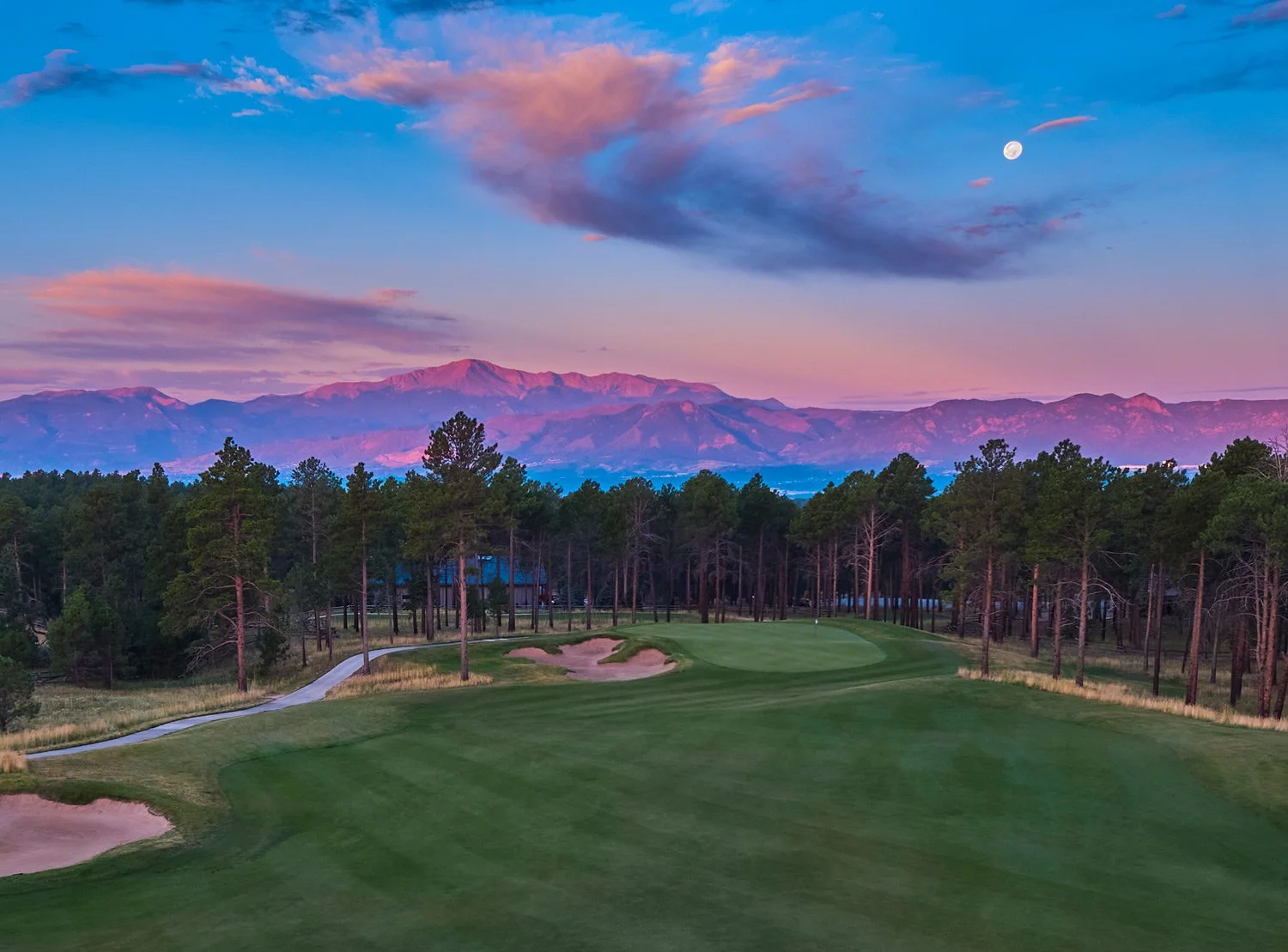 A scenic view of a golf course at sunset with green grass, sand bunkers, tall trees, and a mountain range in the background, under a sky with pink, blue, and purple hues, a full moon visible.