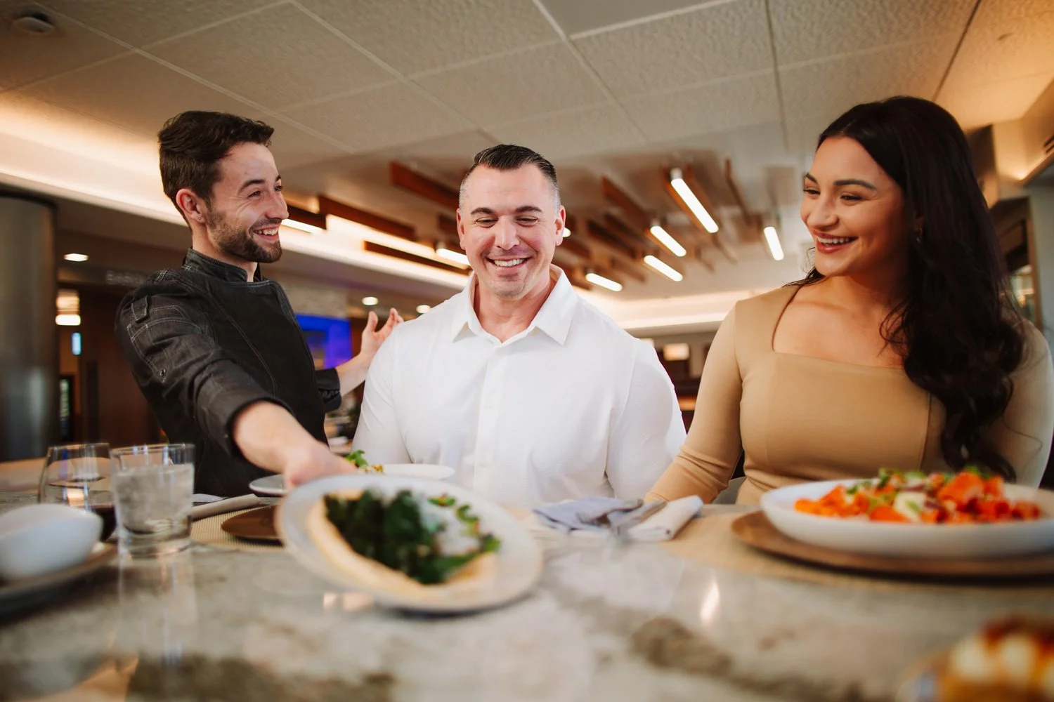 Three people smiling and sharing a meal together in a restaurant, with dishes of food and drinks on the table.