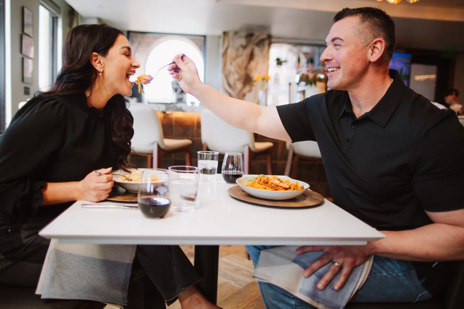 A man feeding a woman pasta in a restaurant, both smiling, with glasses of red wine and water on the table.
