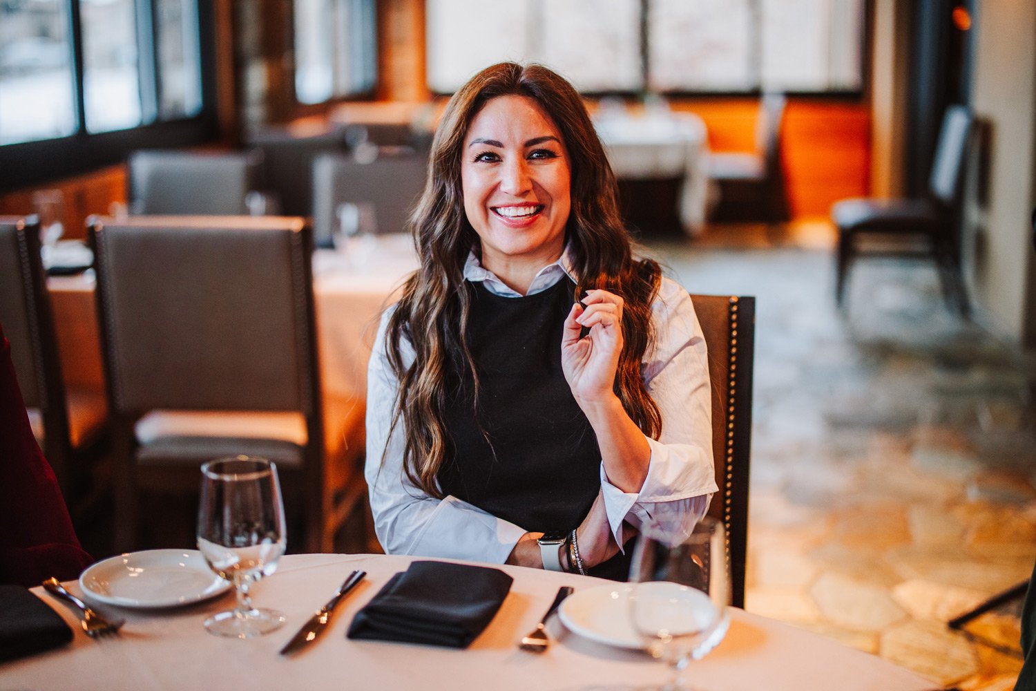 A woman sitting at a restaurant table, smiling, wearing a white shirt with a black vest, in a warmly lit dining area.