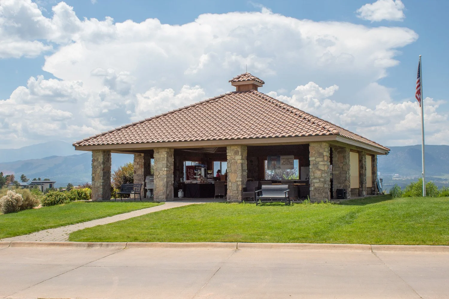 A stone pavilion with a tiled roof sits in a grassy field under blue sky with clouds, with mountains in the background and an American flag flying on a flagpole to the right.