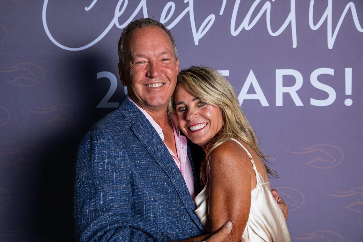 A smiling middle-aged man and woman embracing at a celebration event with a backdrop that reads 'Celebrating 20 Years!' in purple and white lettering.