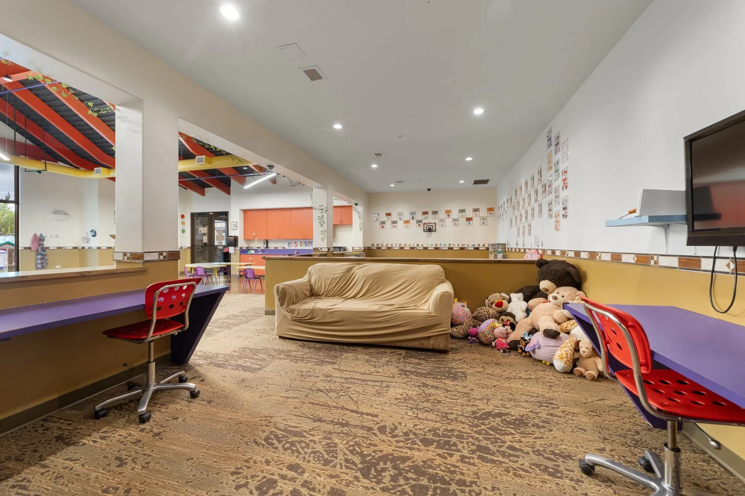Empty indoor children's playroom in the Kids Cabin with purple desks, red chairs, beige couch, toys on the floor, decorated walls, and a large TV on the right.