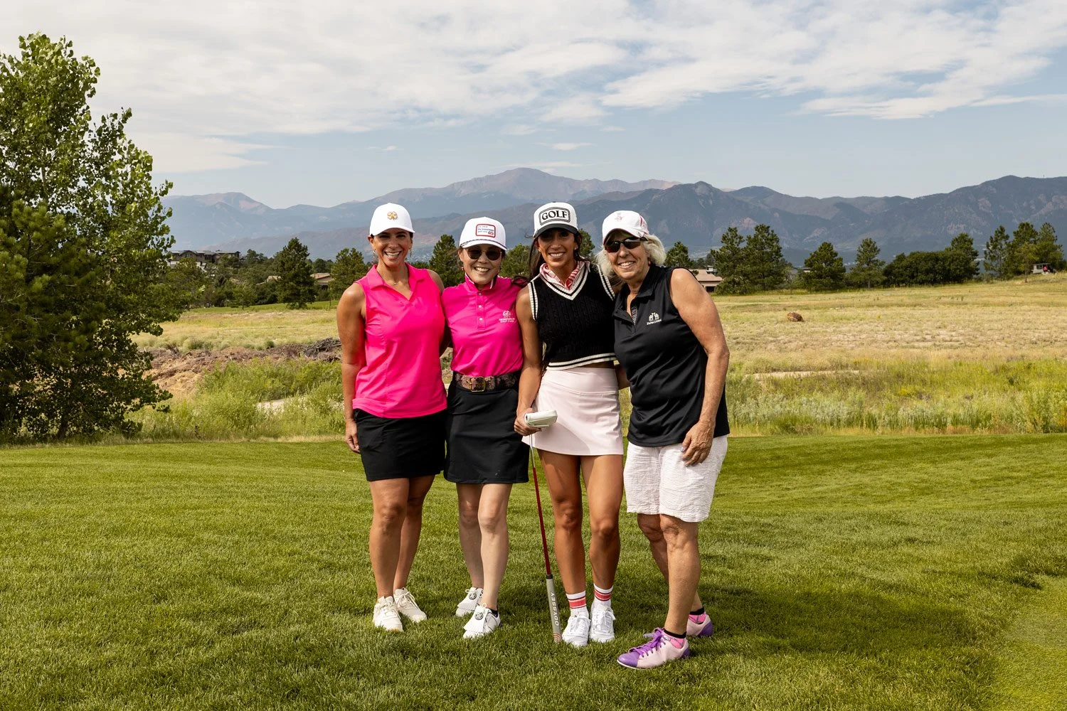 Four women in golf attire standing on a golf course with mountains in the background, smiling at the camera.