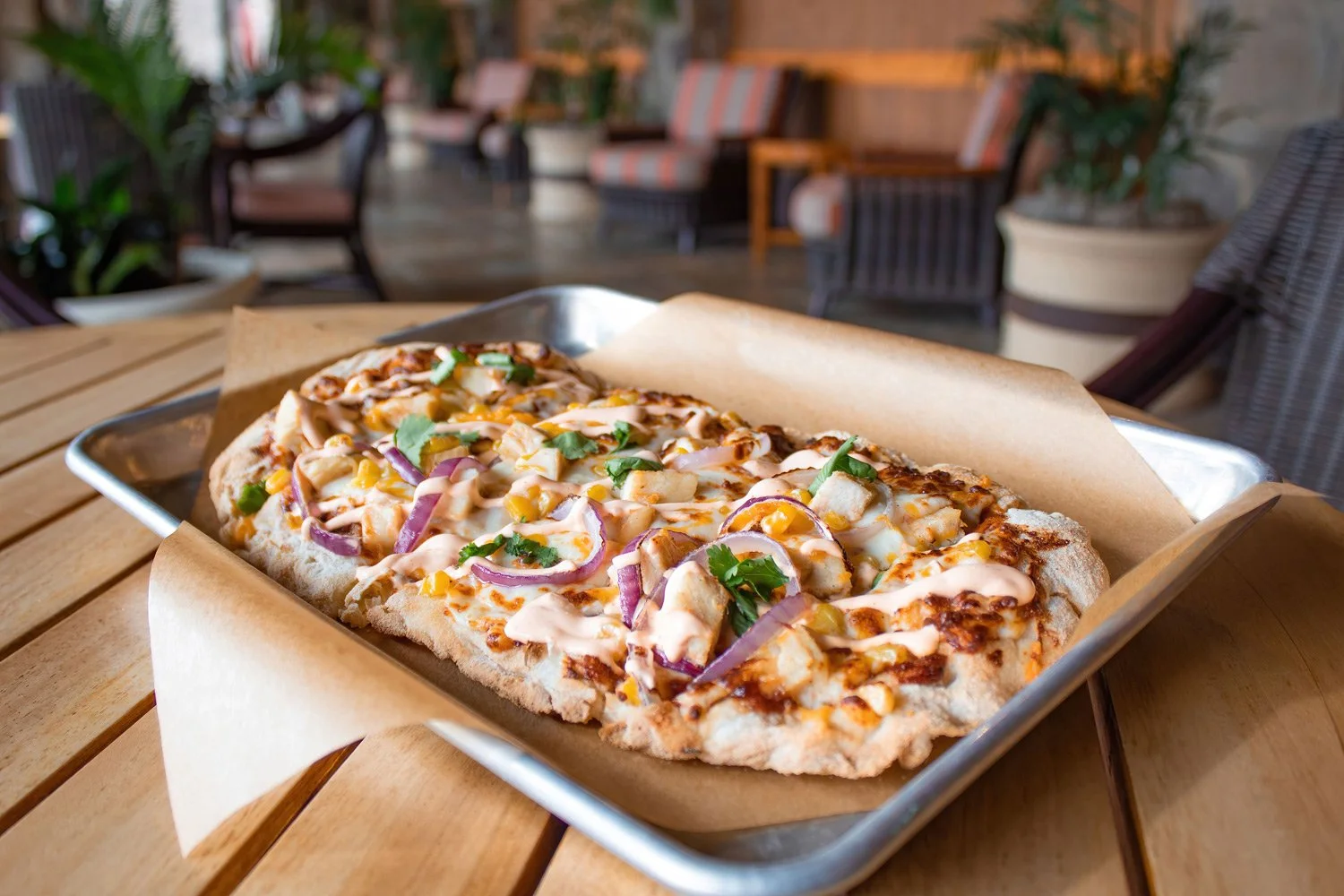 A rectangular pizza with toppings of onion, chicken, corn, and herbs on a metal tray with parchment paper, on a wooden table in a cozy restaurant setting.
