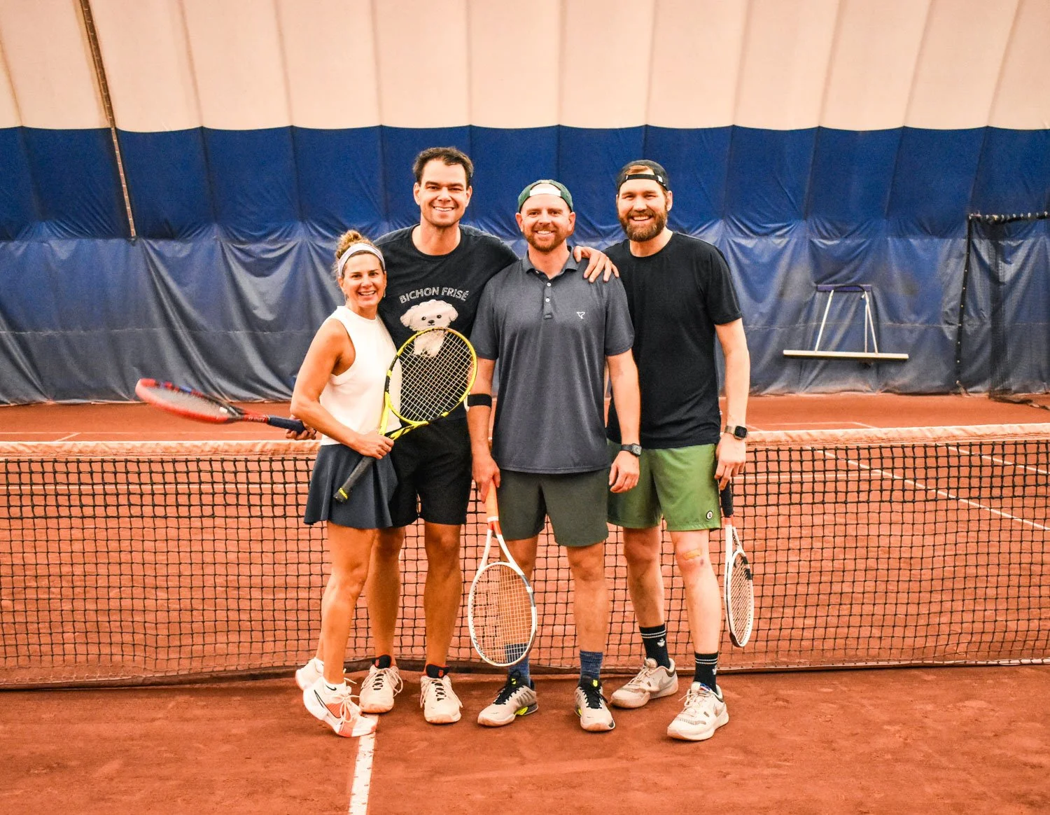 Four people standing on a tennis court, smiling, holding tennis rackets, with a net in front of them and an indoor tennis facility in the background.