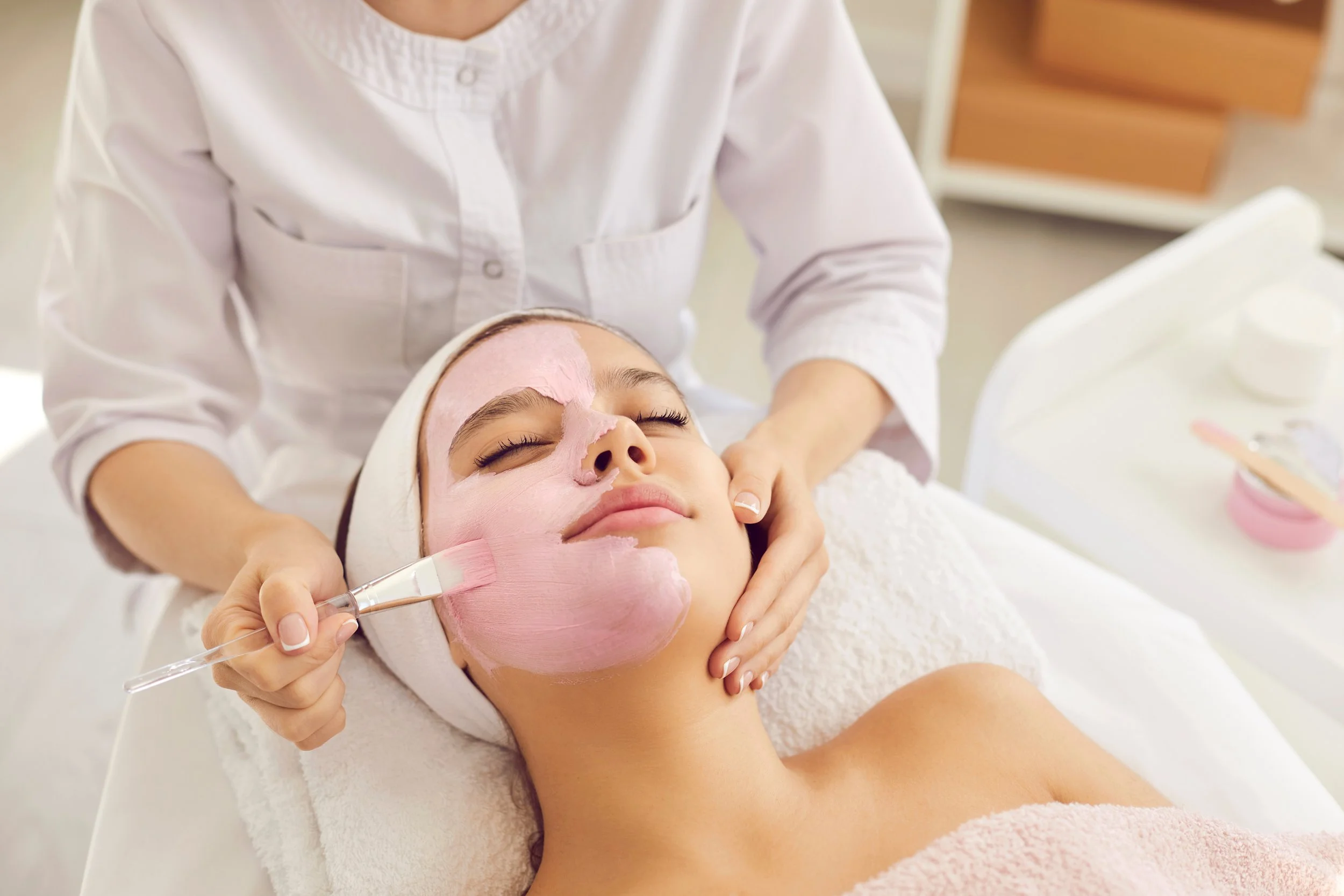 A woman receives a facial treatment, with a beauty professional applying a pink mask to her face, in a spa or skincare clinic.