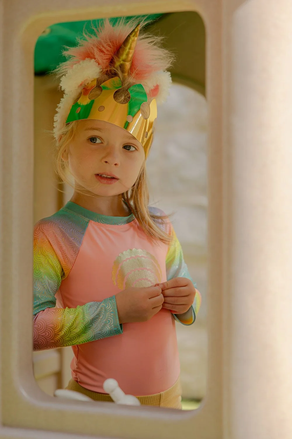 A young girl with blonde hair wearing a gold unicorn horn crown with pink and white fluffy details and a rainbow-colored paper crown on her head, looking through a playhouse window.