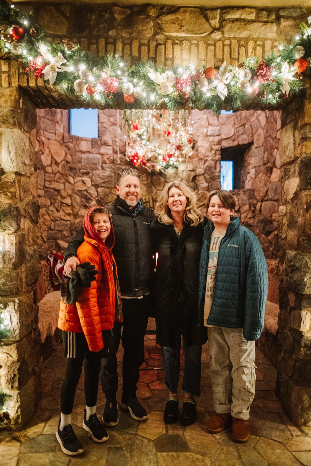 Family of four standing inside a decorated Christmas stone fireplace with holiday ornaments and wreaths.