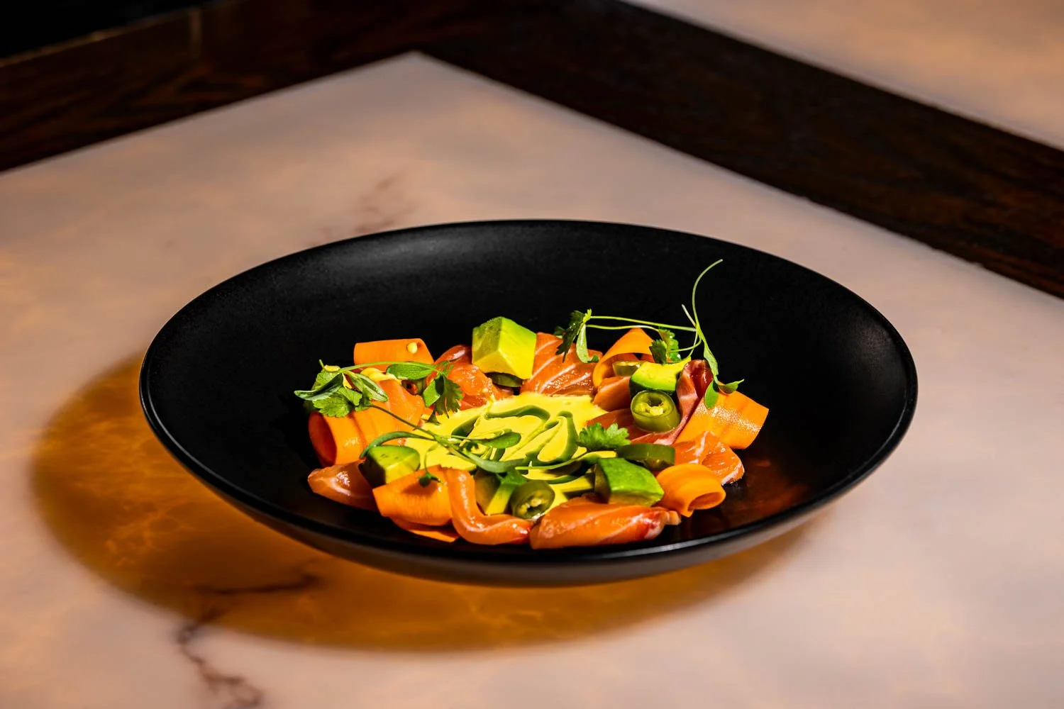 Plate of smoked salmon with avocado, carrots, sliced jalapeños, and microgreens on a black plate.