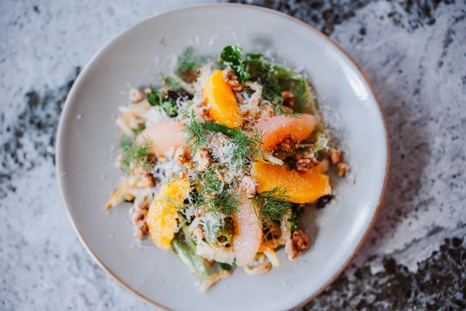 Salad with sliced oranges, greens, grated cheese, chopped nuts, and herbs on a white plate.