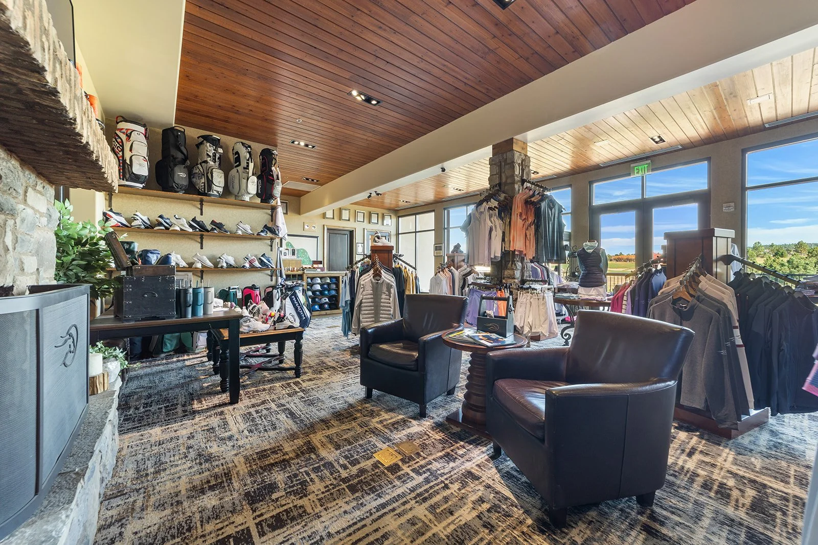 Inside a golf shop with golf bags on the wall, golf shoes on shelves, and clothing racks with golf apparel. There are two black chairs and a wooden table in the foreground, large windows showing a golf course outside, and a wooden ceiling.