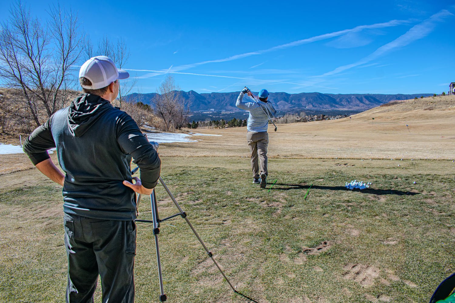 Two men on a golf course: one preparing to hit the ball and the other watching, with mountains in the background under a clear blue sky.
