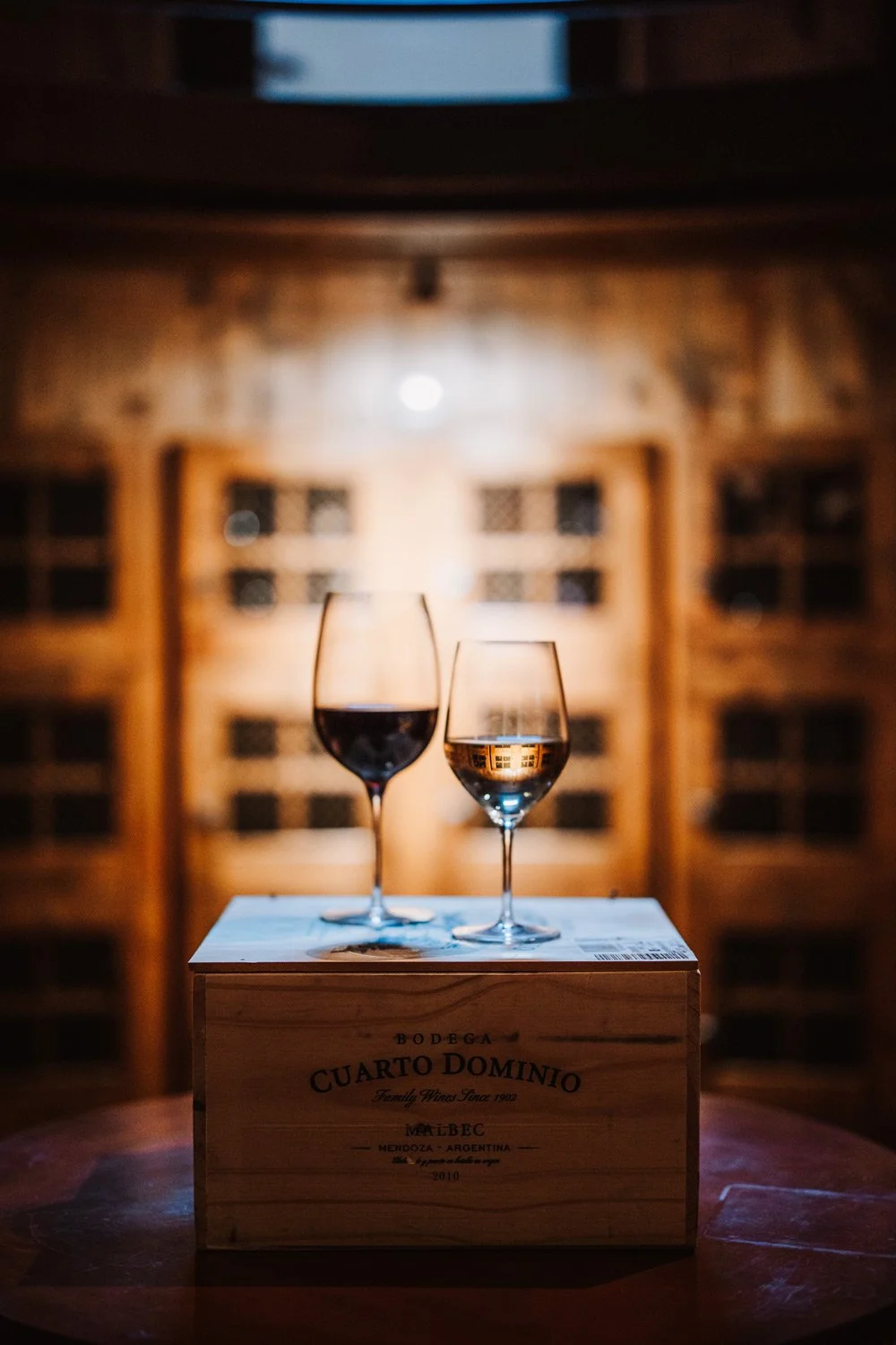 Two glasses of wine, one red and one white, placed on a wooden box inside a warm, rustic wine cellar or winery with wooden shelves in the background.