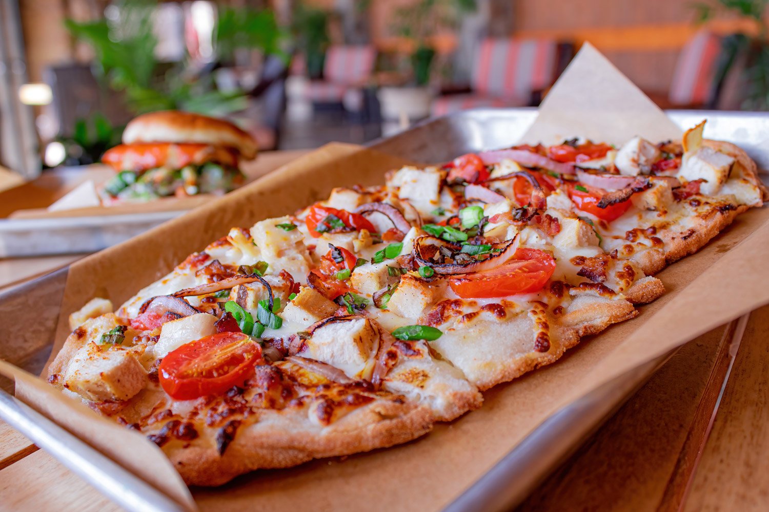 A rectangular pizza with toppings such as cherry tomatoes, green onions, diced chicken, bacon, and sauce, placed on parchment paper in a metal tray, with a sandwich in the background.