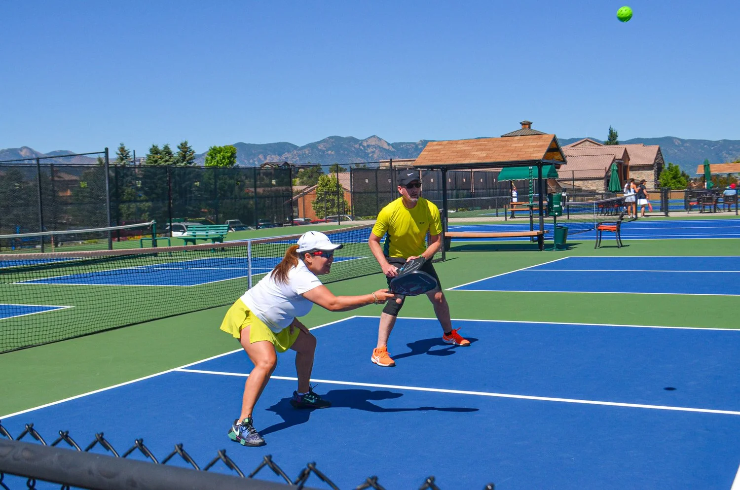 Two people playing pickleball on a sunny outdoor court with a scenic mountain background. The woman in a white shirt, yellow shorts, and white cap is attempting to hit the ball with a paddle, while the man in a yellow shirt, black shorts, and sunglas