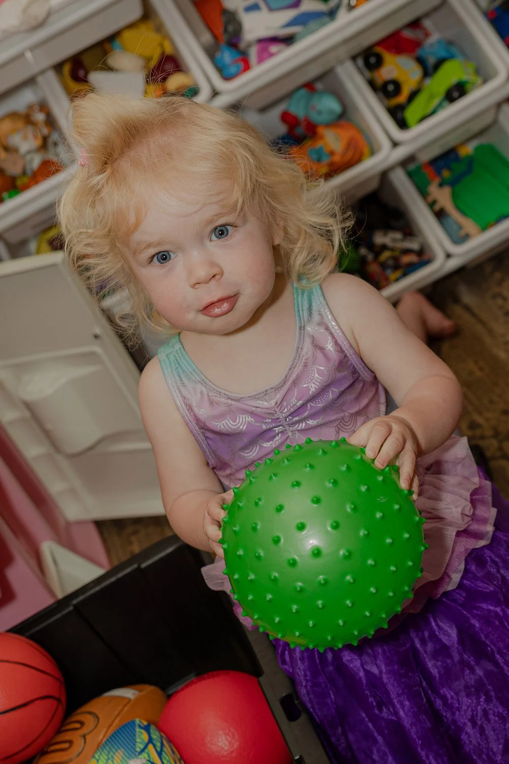 A young girl with curly blonde hair and blue eyes holding a bright green spiky rubber ball inside a toy store.