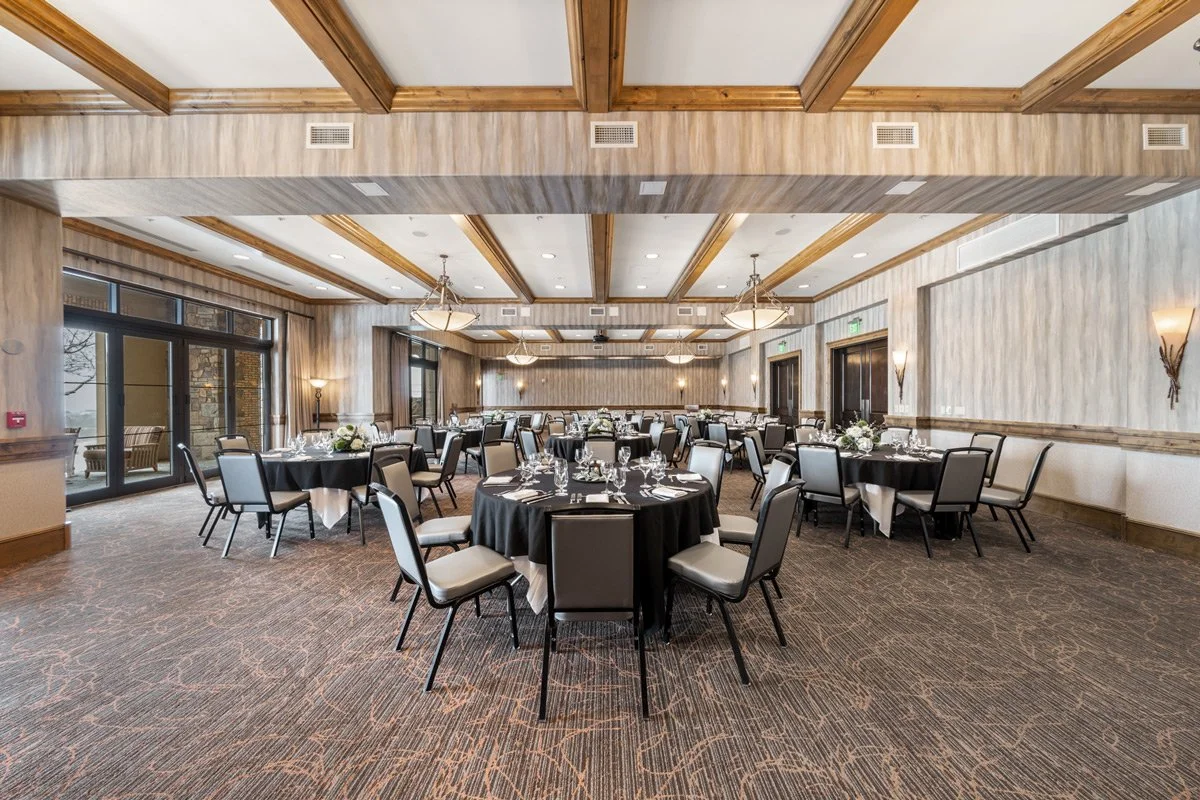 A spacious banquet room with round tables covered in black tablecloths, set with plates, glasses, and silverware, ready for an event.