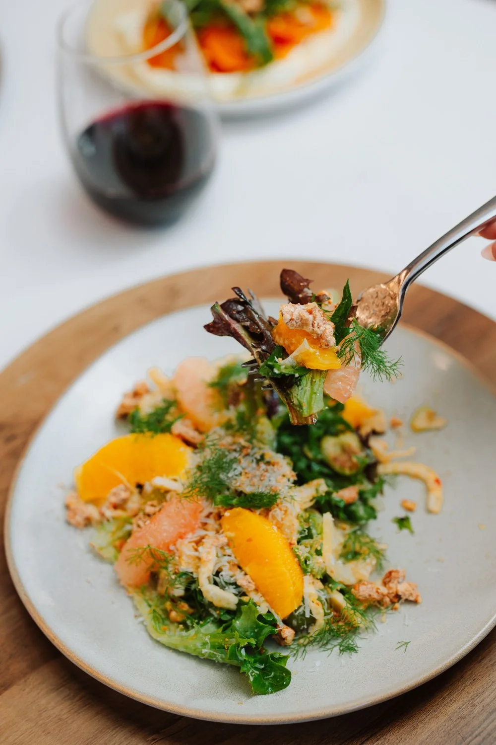 A fork holding a bite of salad above a plate of leafy greens, peach slices, shredded cheese, and herbs, with a glass of red wine in the background.