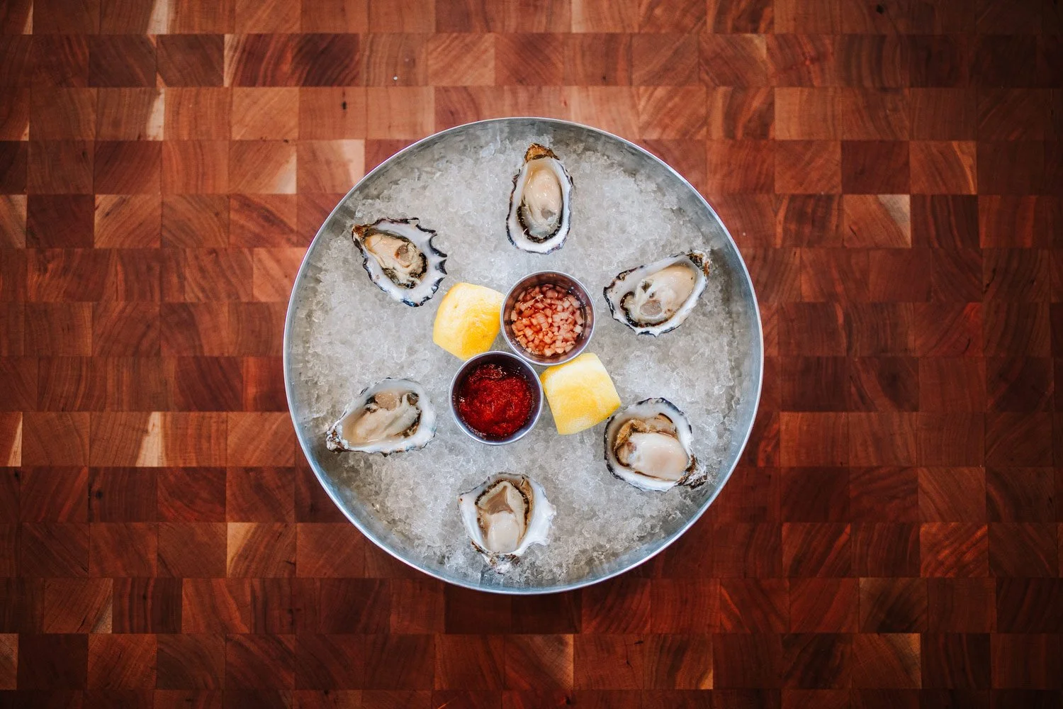 A round metal tray with ice, holding six oysters on the half shell, lemon wedges, and small cups of cocktail sauce and hot sauce. The tray is on a wooden surface.