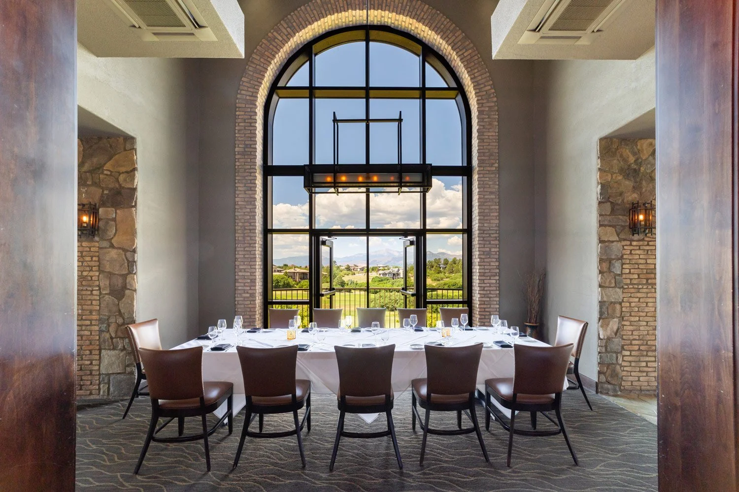 A dining room with large arched window overlooking a scenic landscape, a long table set for a meal with chairs around it, and a modern chandelier hanging above at The Steakhouse.