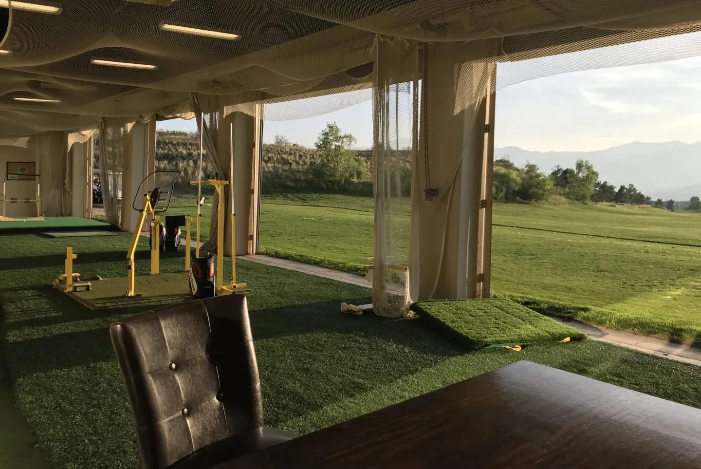 Indoor golf practice facility with artificial turf, exercise equipment, and large open windows opening to a scenic outdoor view of green grass, trees, and distant mountains at sunset.