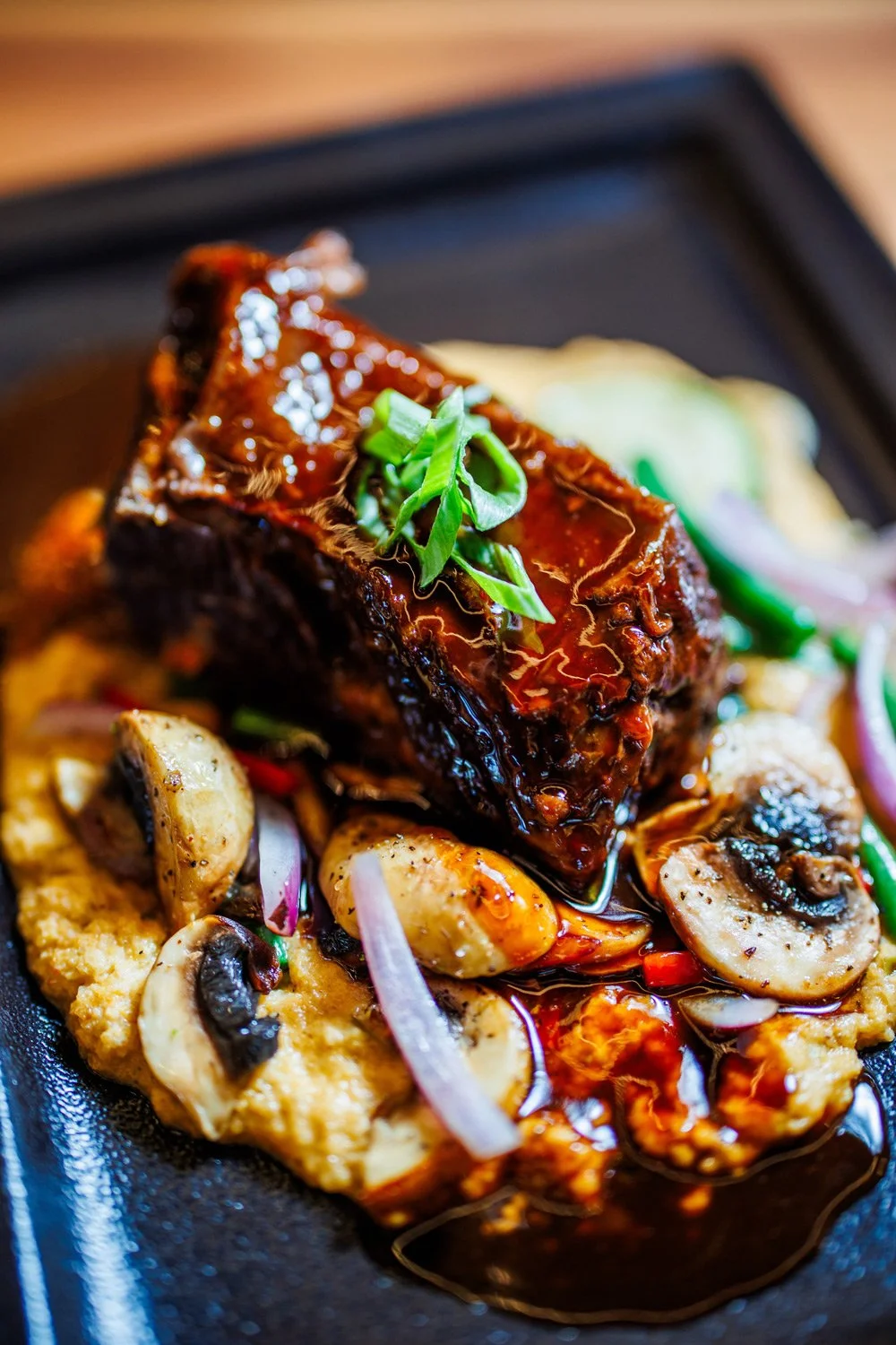 A plate of glazed short ribs served with sautéed mushrooms, onions, and a bed of mashed potatoes, garnished with chopped green onions.