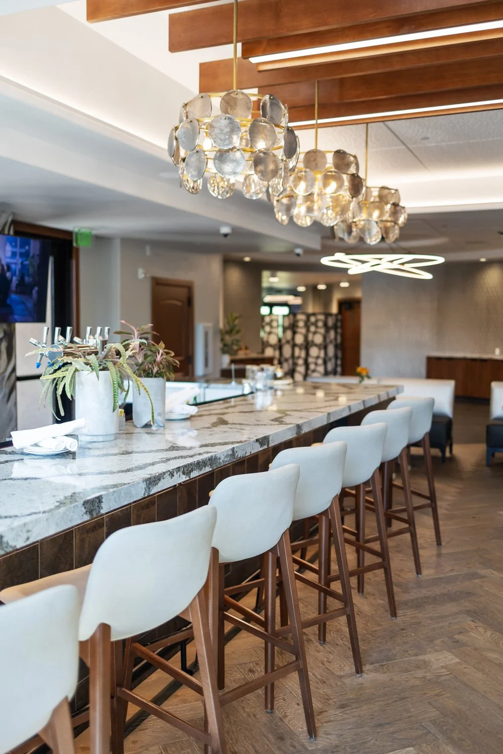 A modern hotel bar with a marble countertop, white upholstered bar stools with wooden legs, hanging decorative lights, and a cozy seating area in the background.