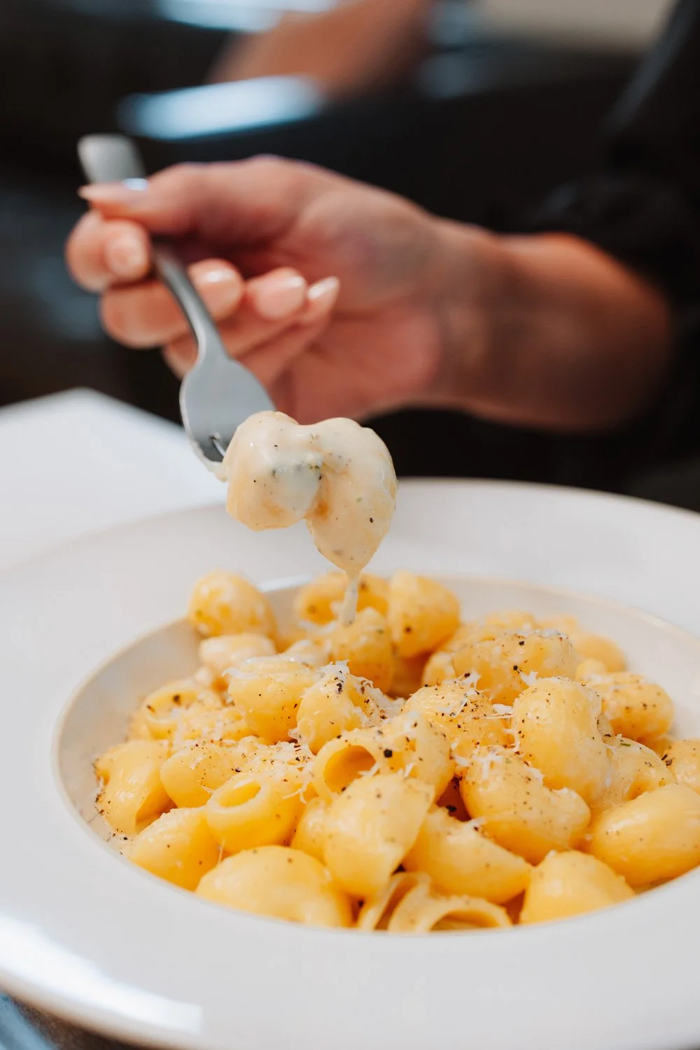Someone holding a fork with a piece of creamy cheese over a white plate of cooked pasta noodles topped with grated cheese and black pepper.