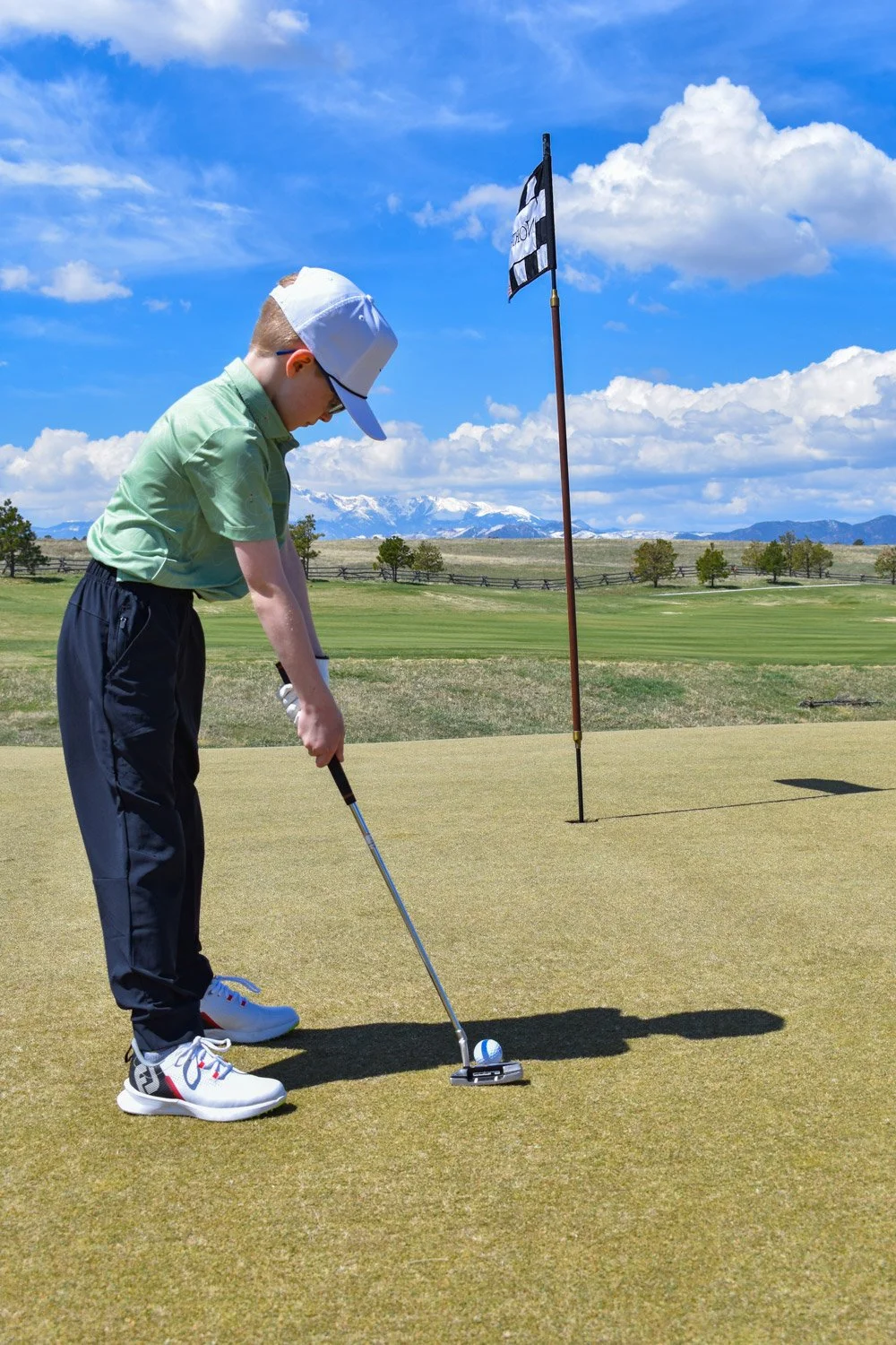 Young boy preparing to putt on a golf course with mountains in the background and a flag marked 'GOLF' nearby.