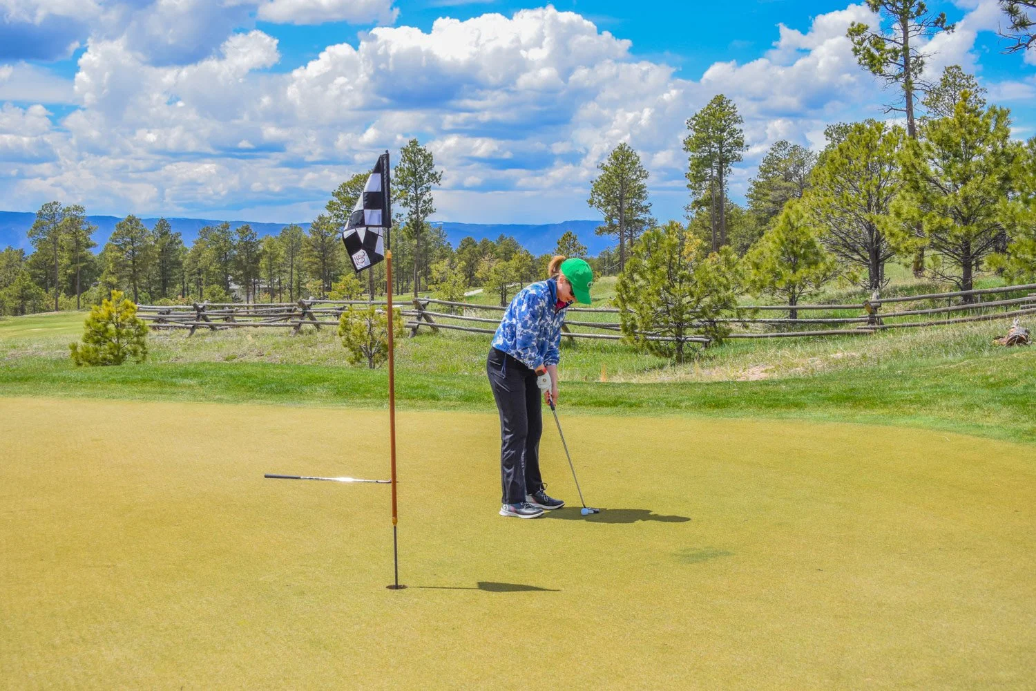 A woman in a blue patterned jacket and green cap playing golf on a green putting surface surrounded by trees and a rural wooden fence under a partly cloudy sky
