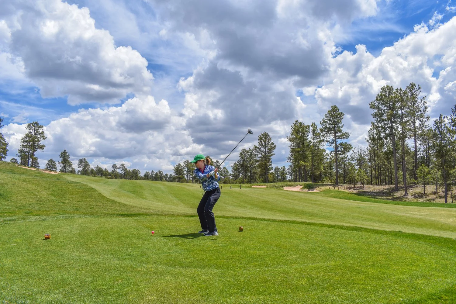 A woman playing golf on a bright, partly cloudy day. She is preparing to hit a pink golf ball on the tee box, surrounded by lush green grass and a backdrop of tall trees and open sky.
