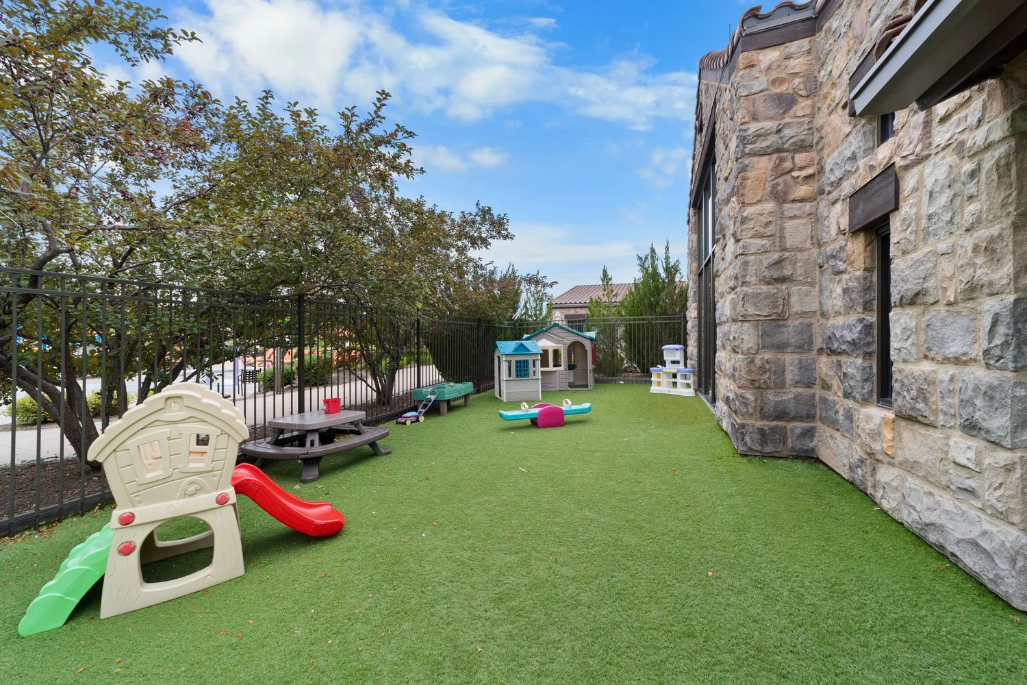 Kids' Cabin backyard with playset, sandbox, small playhouses, and toy boat on artificial grass, enclosed by a black fence, with a stone house wall on the right and trees on the left under a partly cloudy sky.