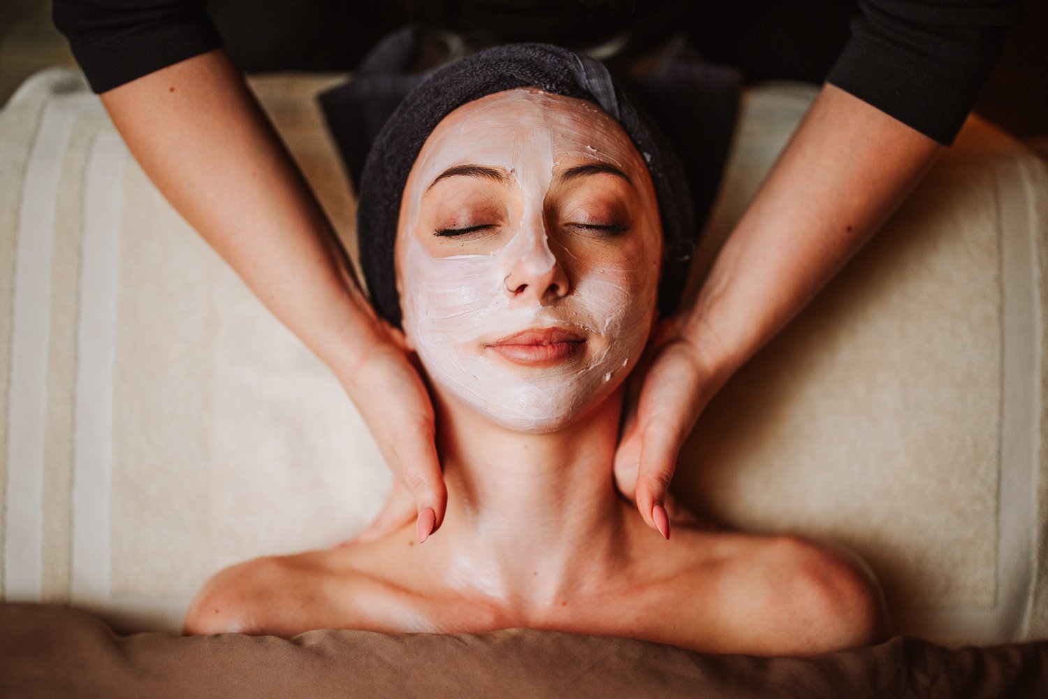 Woman receiving a facial massage at a spa, with a facial mask applied on her face, lying on a massage bed with a towel wrapped around her head.