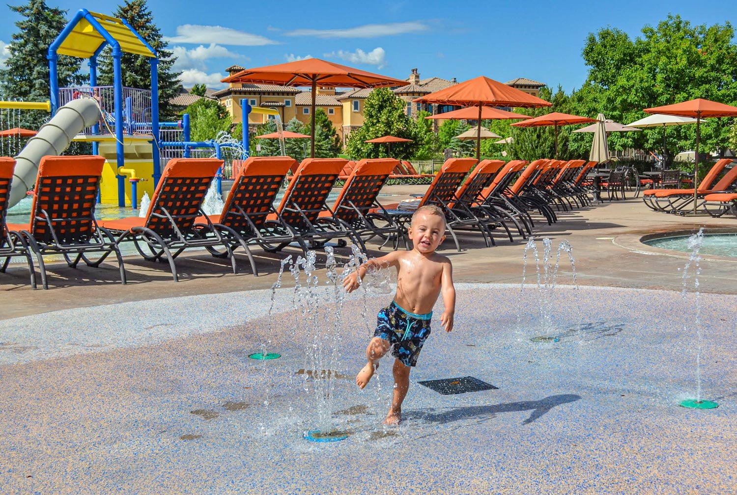 A young boy playing in a splash pad area with water fountains, surrounded by pool chairs and lounge umbrellas on a sunny day.