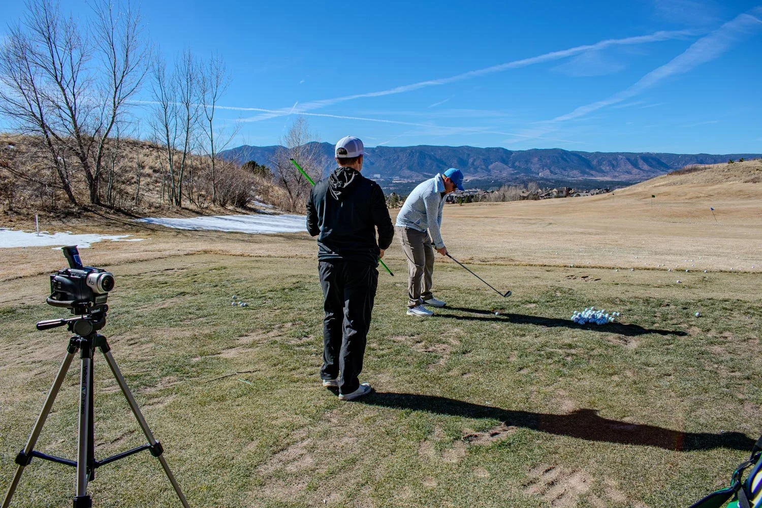 Two men on a golf course practicing their swings. One is preparing to hit a golf ball, while the other is observing. There is a camera on a tripod set up nearby.