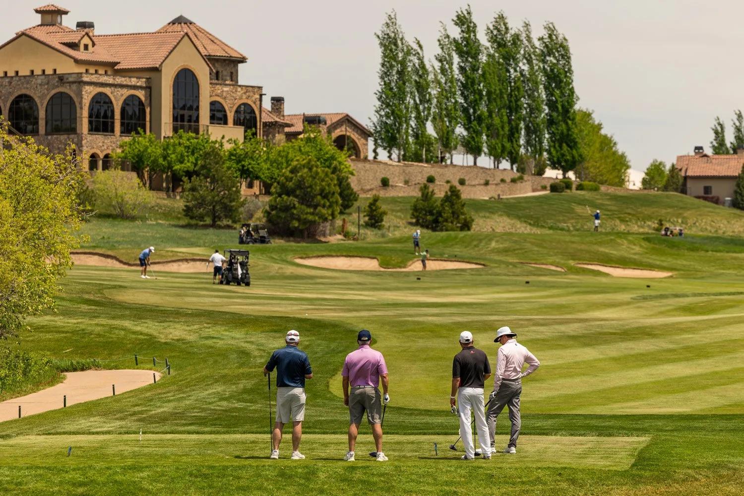A group of four men on a golf course, preparing to hit a golf ball, with multiple players and golf carts in the background, outside a The Steakhouse with a terracotta roof and arched windows.