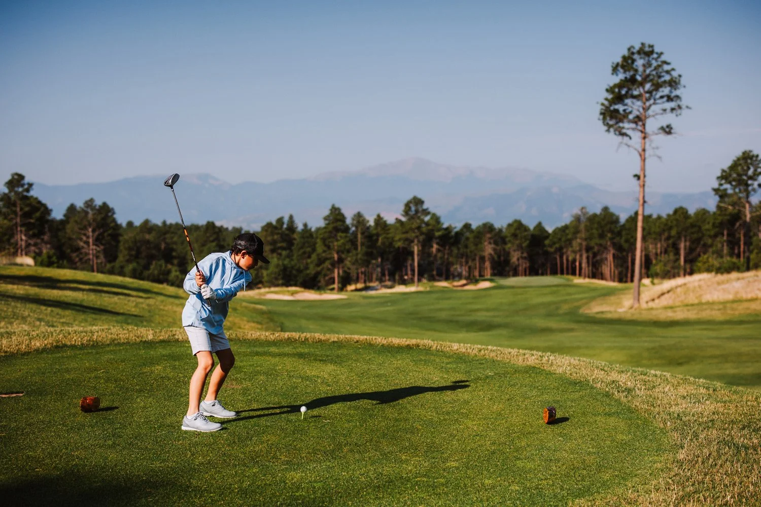 A young boy in athletic clothing wearing a baseball cap, glasses, and sneakers is preparing to hit a golf ball on a golf course with trees, grass, and mountains in the background on a clear day.