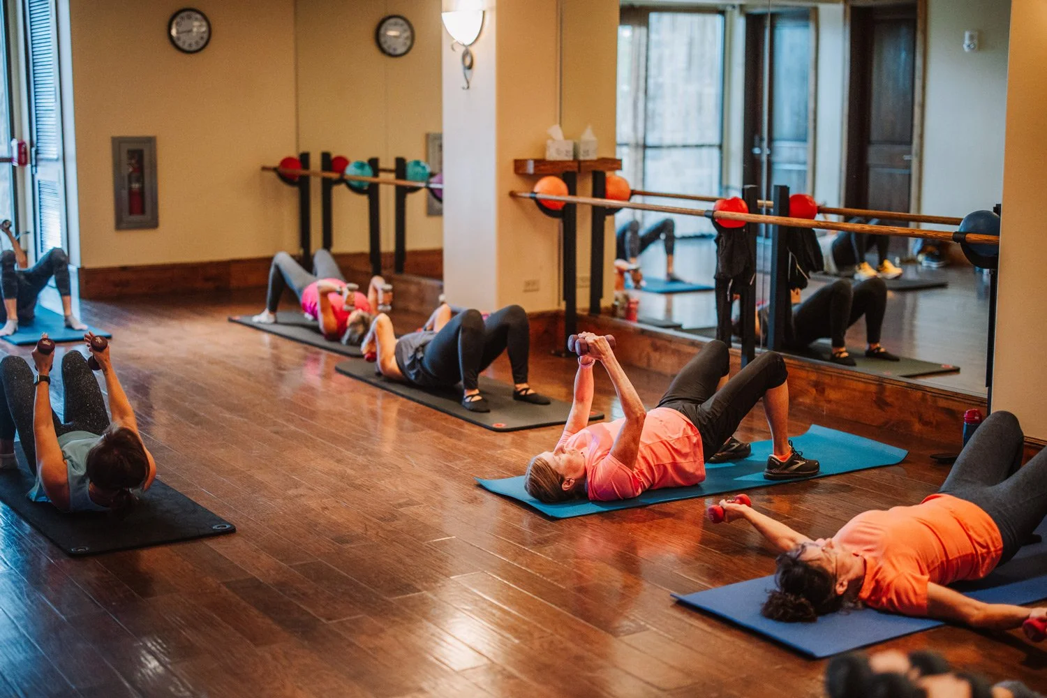Group of seniors participating in a seated exercise class in a fitness studio, lying on exercise mats and holding small red weights, with resistance bands attached to the wall.