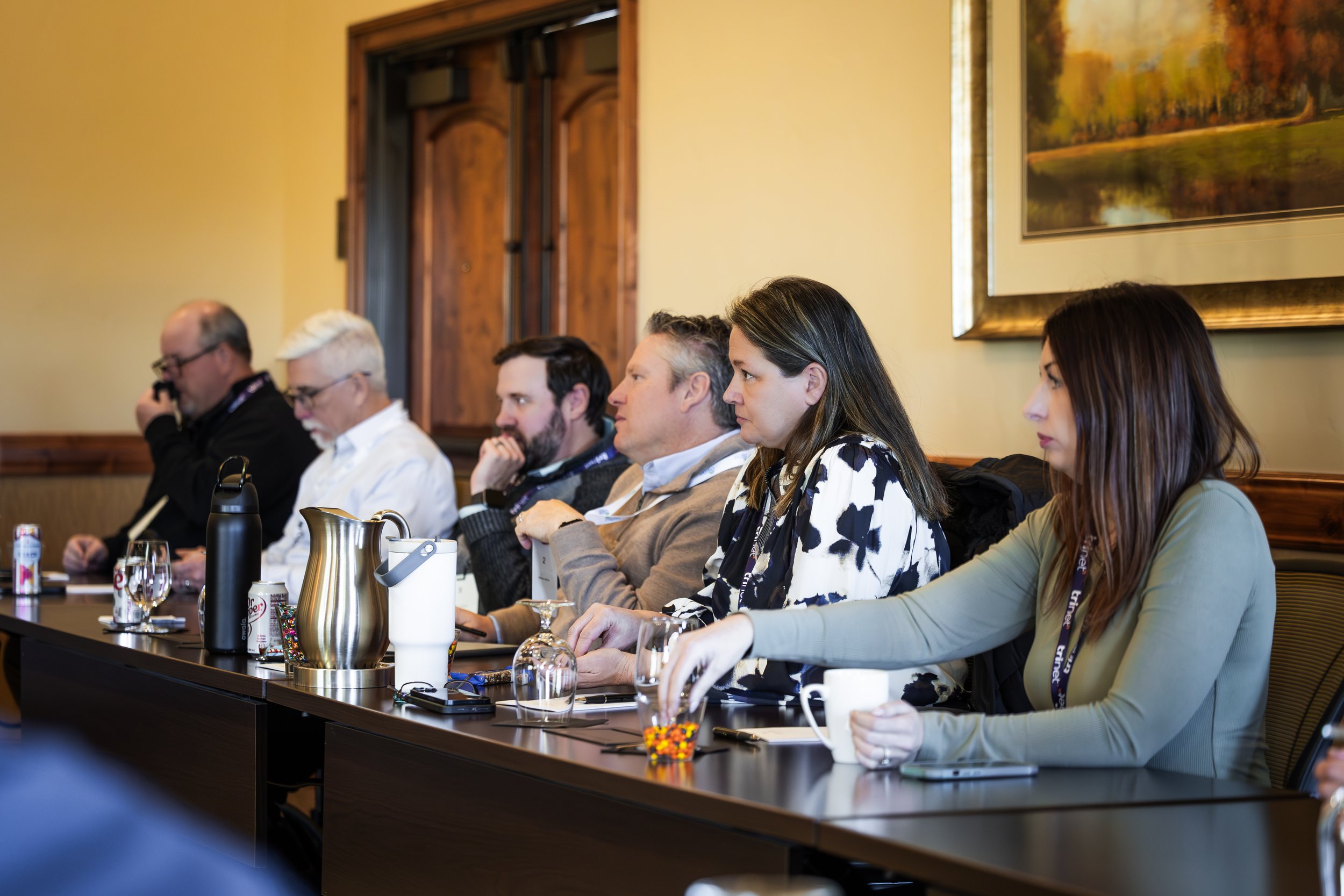Six people sitting at a conference table, some with drinks, attending a meeting or presentation in a room with a large landscape painting on the wall.