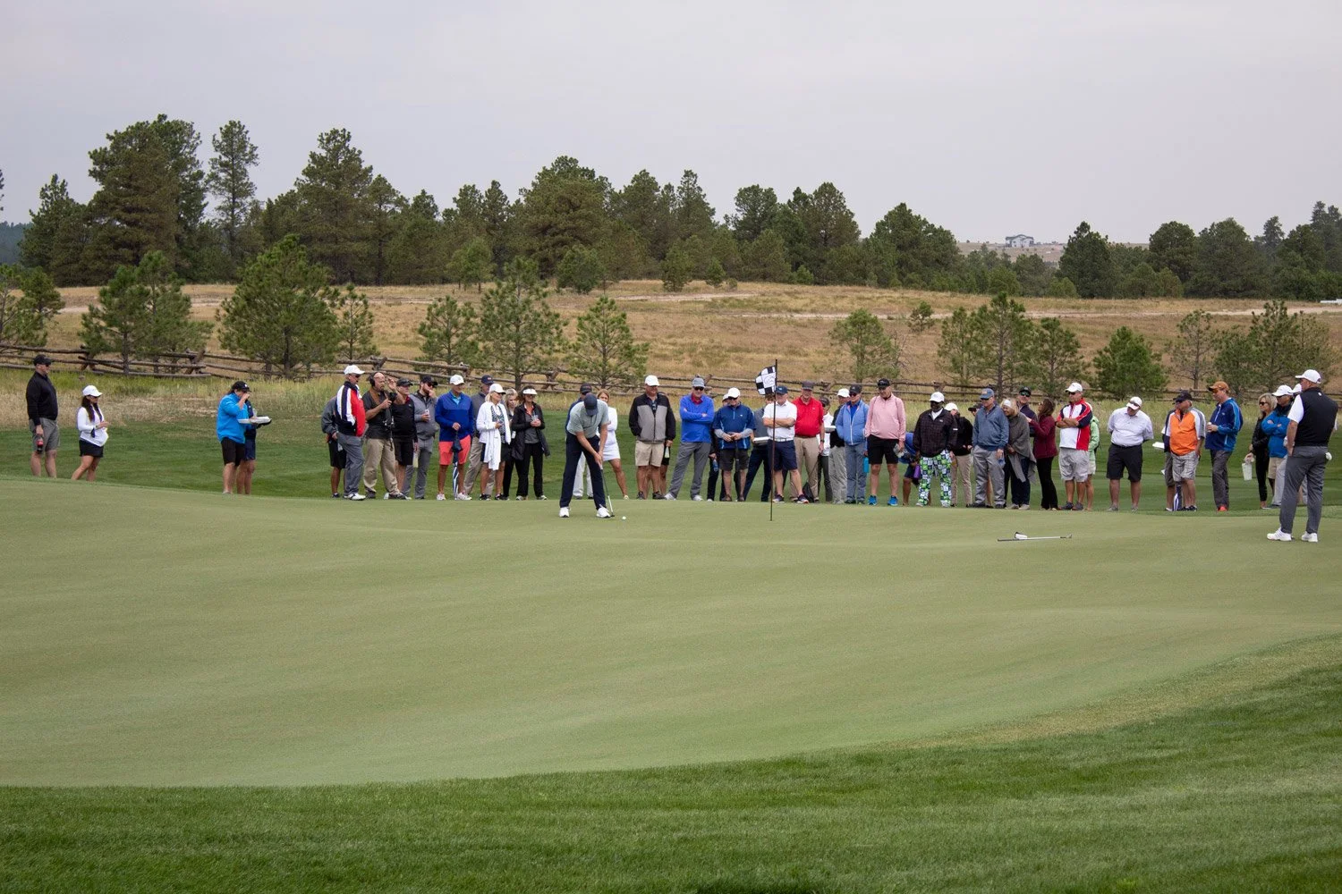 Group of people watching a golfer putt on a golf course. The course has green grass, and trees in the background under a cloudy sky.