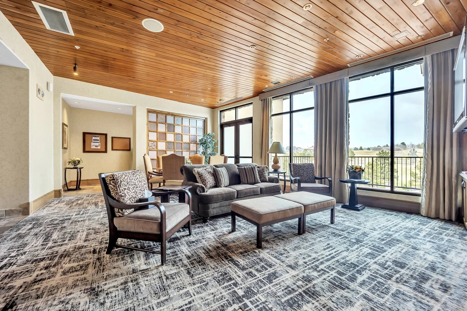 A spacious hotel lounge area with large windows overlooking a green landscape, furnished with a gray sofa, patterned armchairs, a coffee table, side tables with lamps, and a wooden ceiling.
