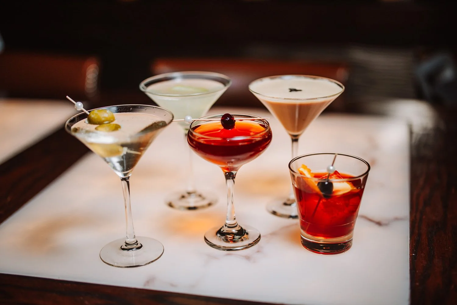 Six cocktails in various glasses arranged on a marble surface, some garnished with olives or fruit slices.