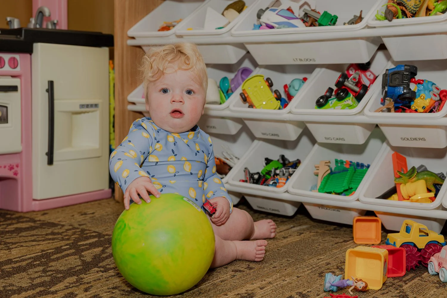 A young child with curly blonde hair sitting on a carpeted floor, holding a green ball and a small toy car, surrounded by assorted toys and a toy kitchen with toy bins filled with various small toys.