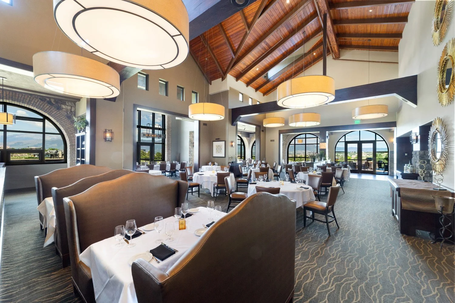 Elegant Steakhouse interior with high vaulted wooden ceiling, large circular pendant lights, arched windows showing a scenic view, and tables set with white tablecloths, wine glasses, and black napkins.