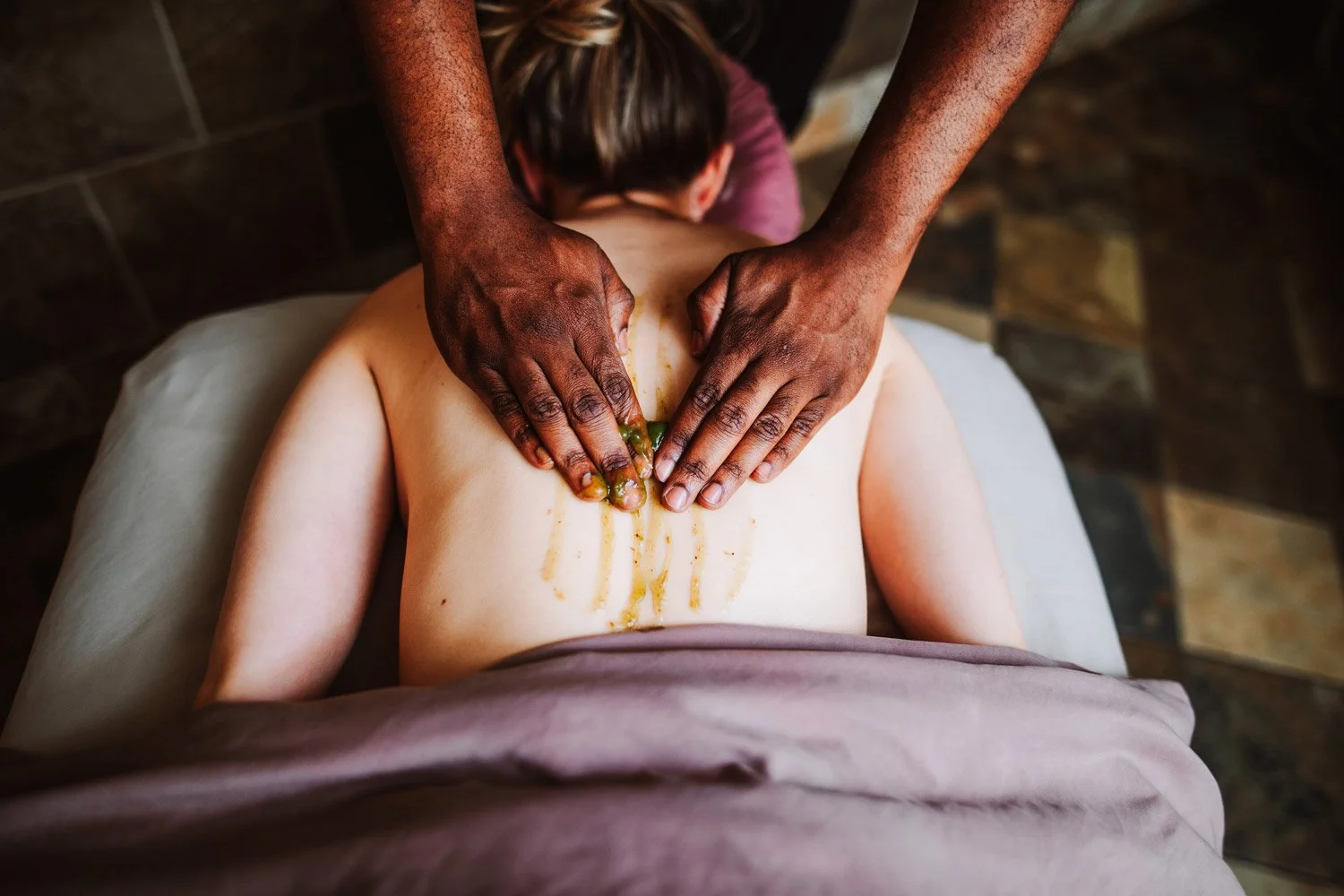 Person giving a back massage to a client lying face down on a massage table, with massage oil and green herb in hands.