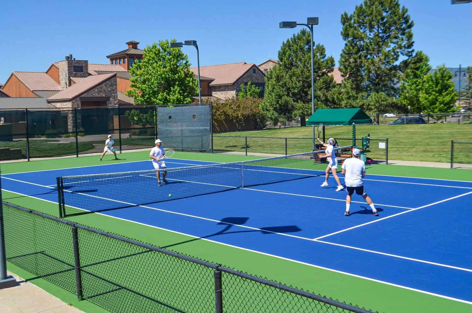 People playing pickleball on a blue court with green borders, surrounded by a black fence, in a sunny outdoor setting with houses, trees, and a park bench in the background.