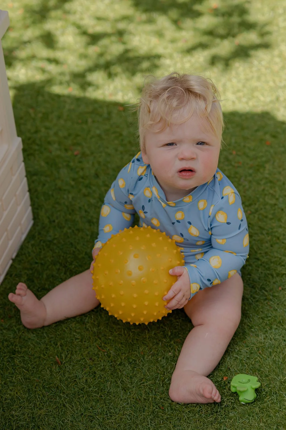 A young child with curly blonde hair sitting on grass, holding a yellow spiky ball, wearing a blue shirt with lemon prints.