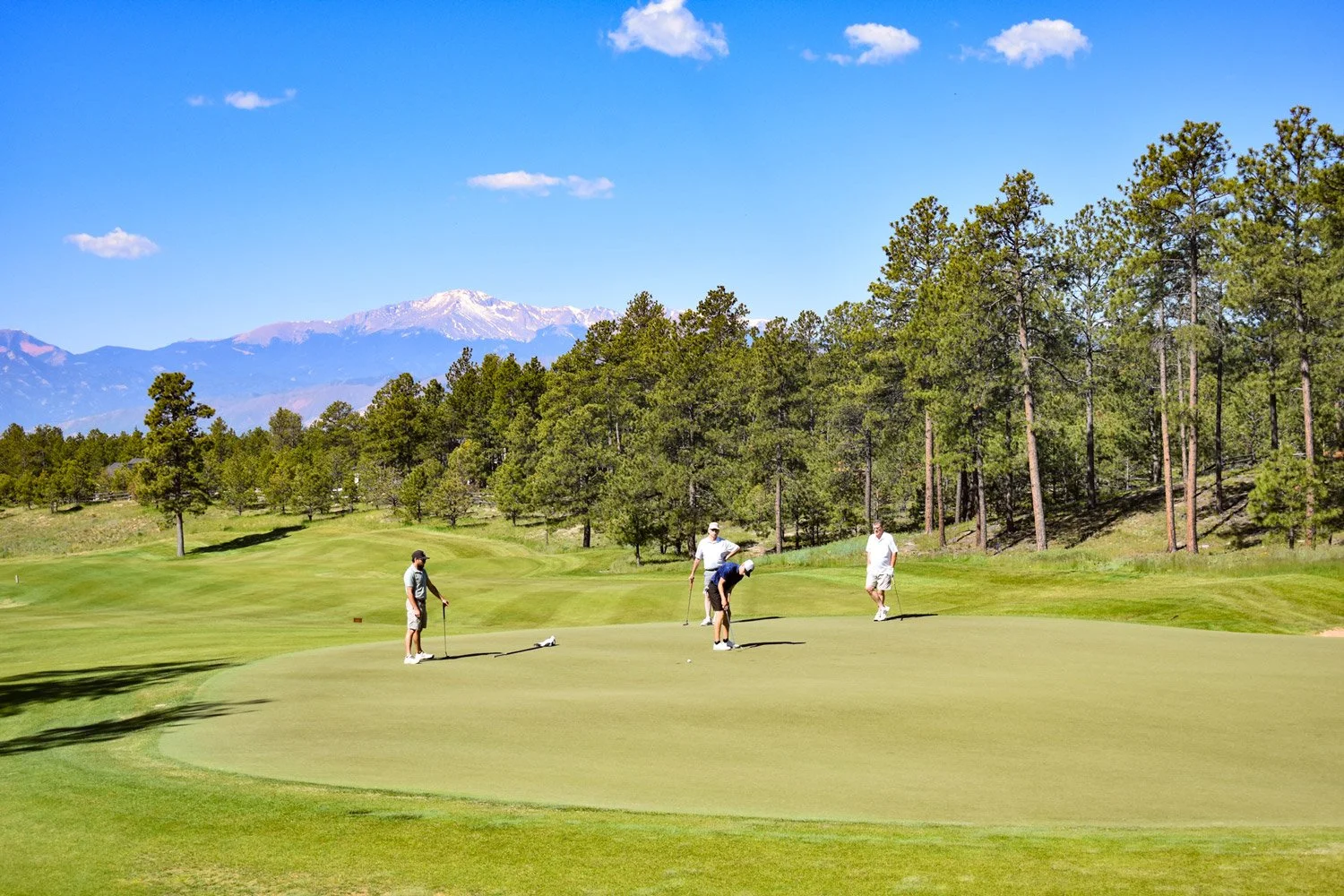 Four people playing golf on a green tee box, with trees and mountains in the background.